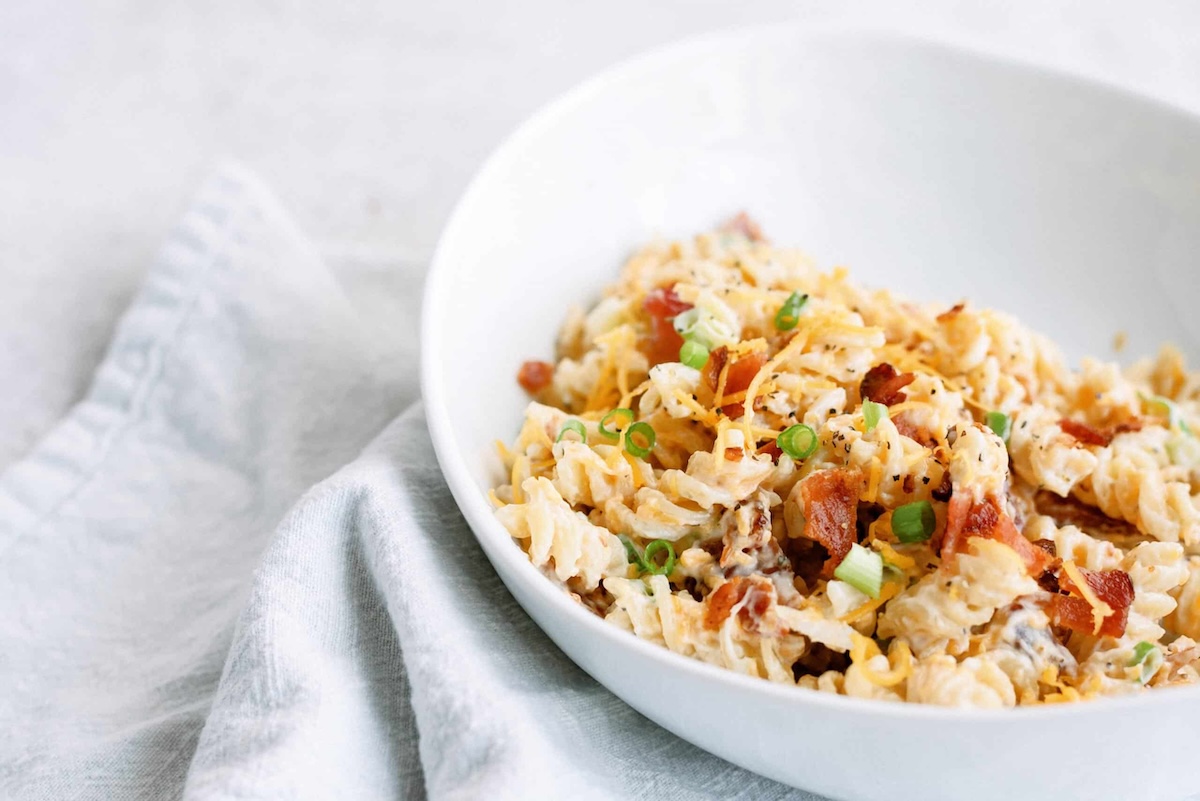 A white bowl filled with pasta salad garnished with chopped green onions and bacon, placed on a light-colored napkin.