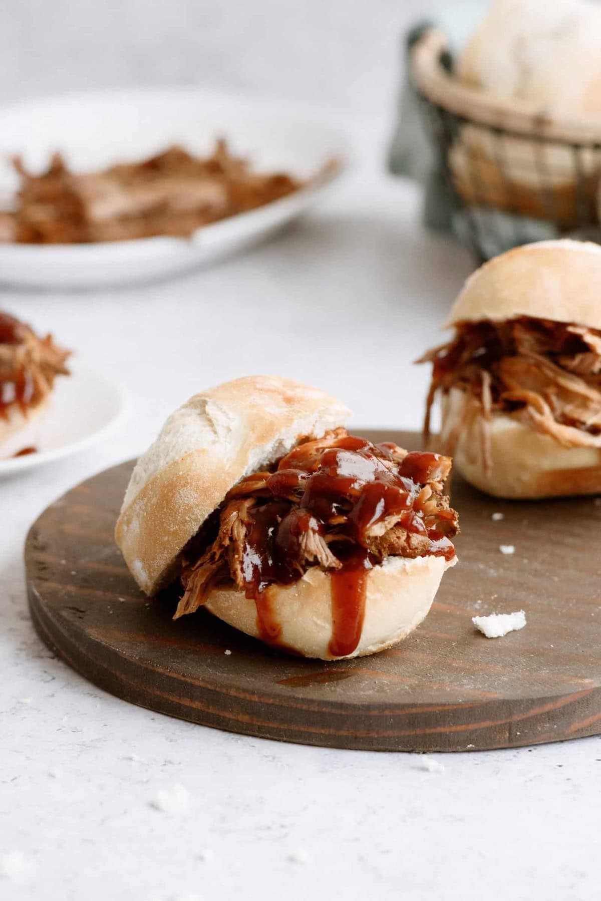 Pulled pork sandwiches topped with barbecue sauce are served on a wooden board, with additional pulled pork and bread visible in the background.