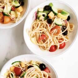 Three white bowls filled with spaghetti pasta salad containing cherry tomatoes, black olives, cucumber, cheese cubes, red onion, and yellow squash on a white surface.