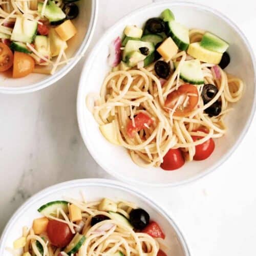 Three white bowls filled with spaghetti pasta salad containing cherry tomatoes, black olives, cucumber, cheese cubes, red onion, and yellow squash on a white surface.