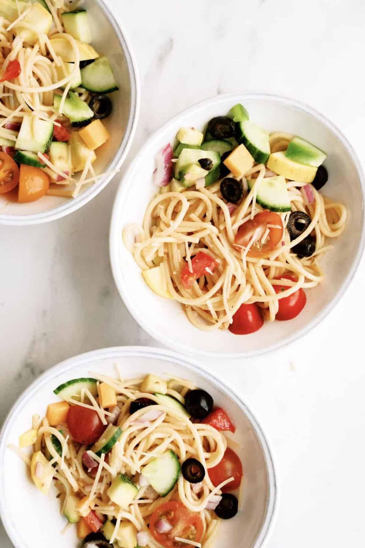 Three white bowls filled with spaghetti pasta salad containing cherry tomatoes, black olives, cucumber, cheese cubes, red onion, and yellow squash on a white surface.