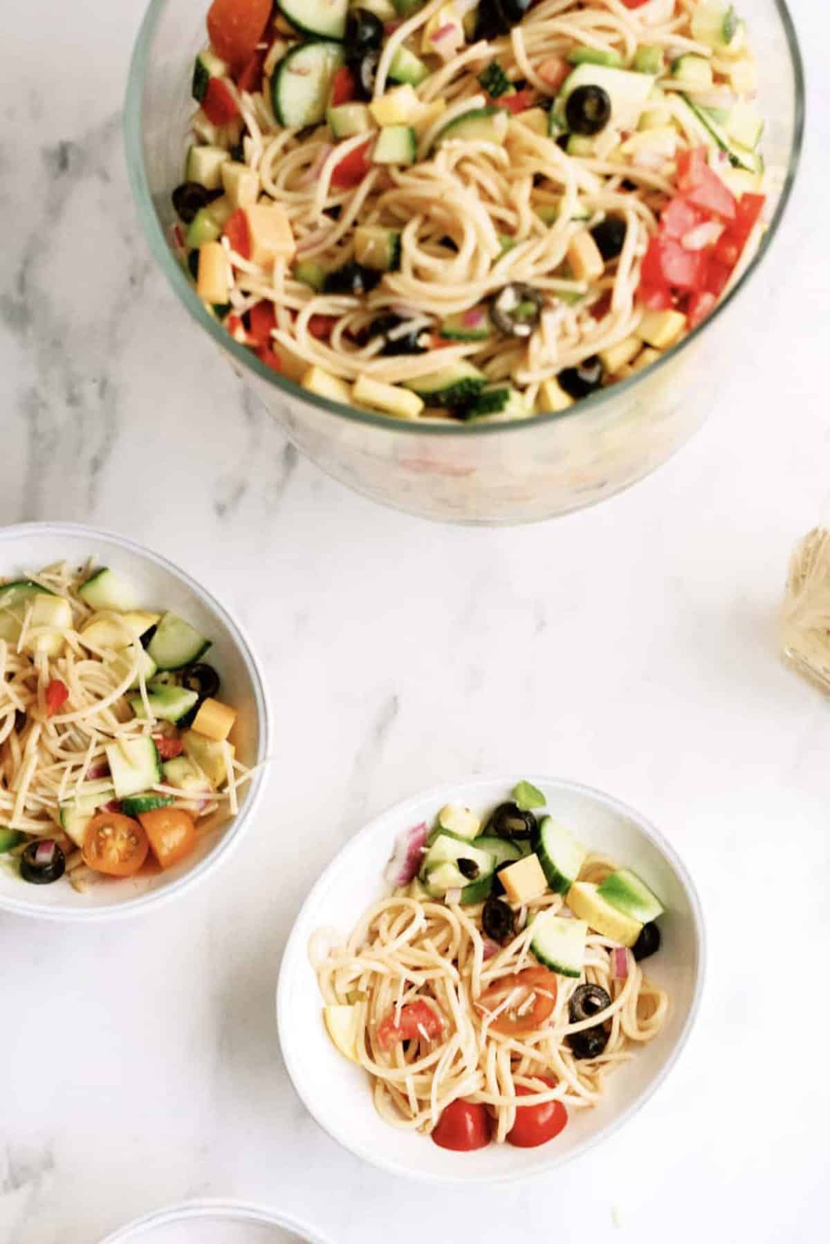 A large glass bowl and two smaller bowls filled with spaghetti pasta salad containing tomatoes, olives, cheese, cucumbers, and other vegetables on a white surface.