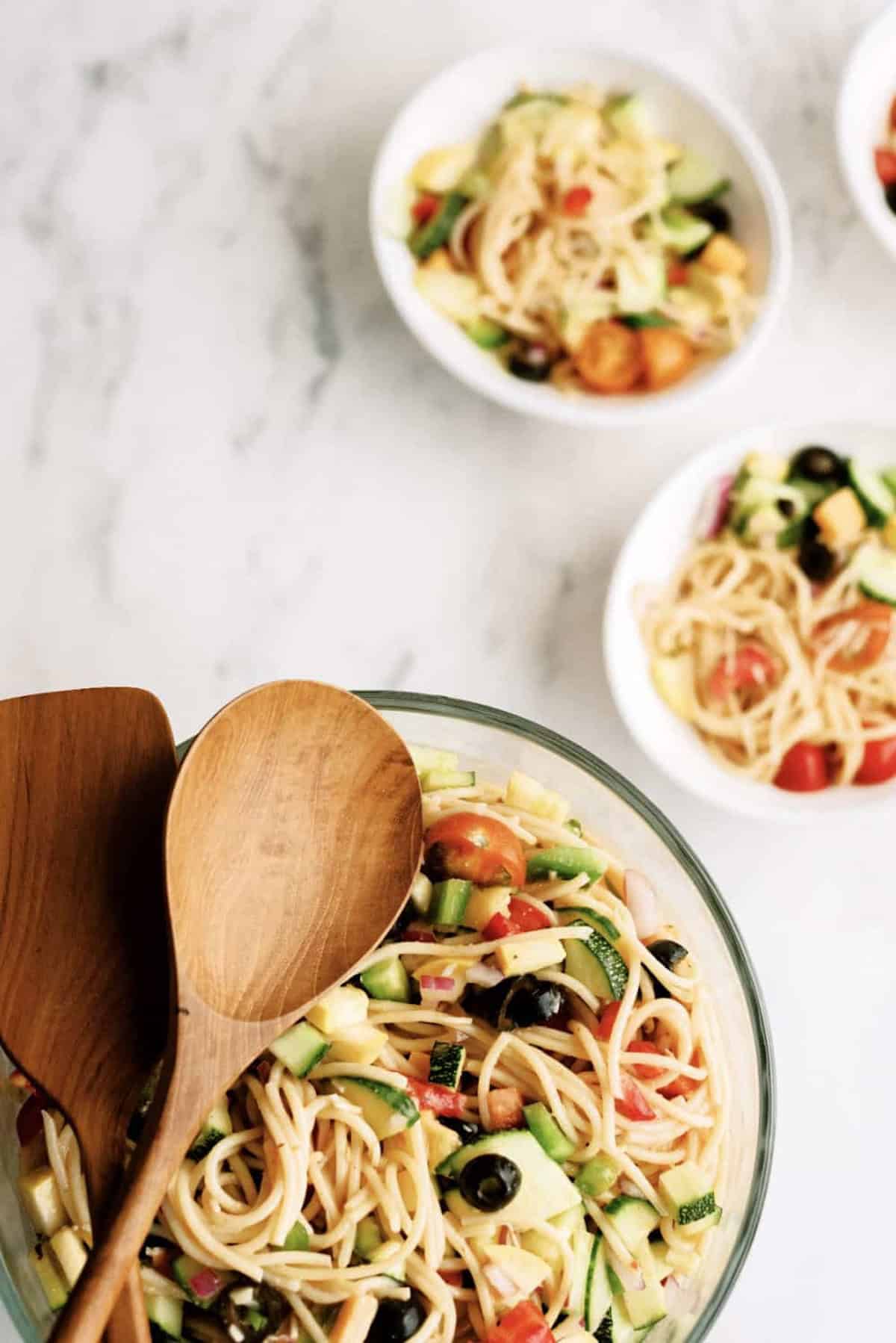 A glass bowl of pasta salad with vegetables and wooden serving utensils next to three small bowls each containing a serving of the same pasta salad.