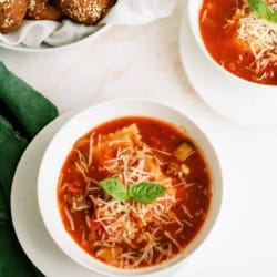 Two bowls of tomato-based soup topped with shredded cheese and basil sit on white plates next to a green napkin, with seeded bread rolls in the background.