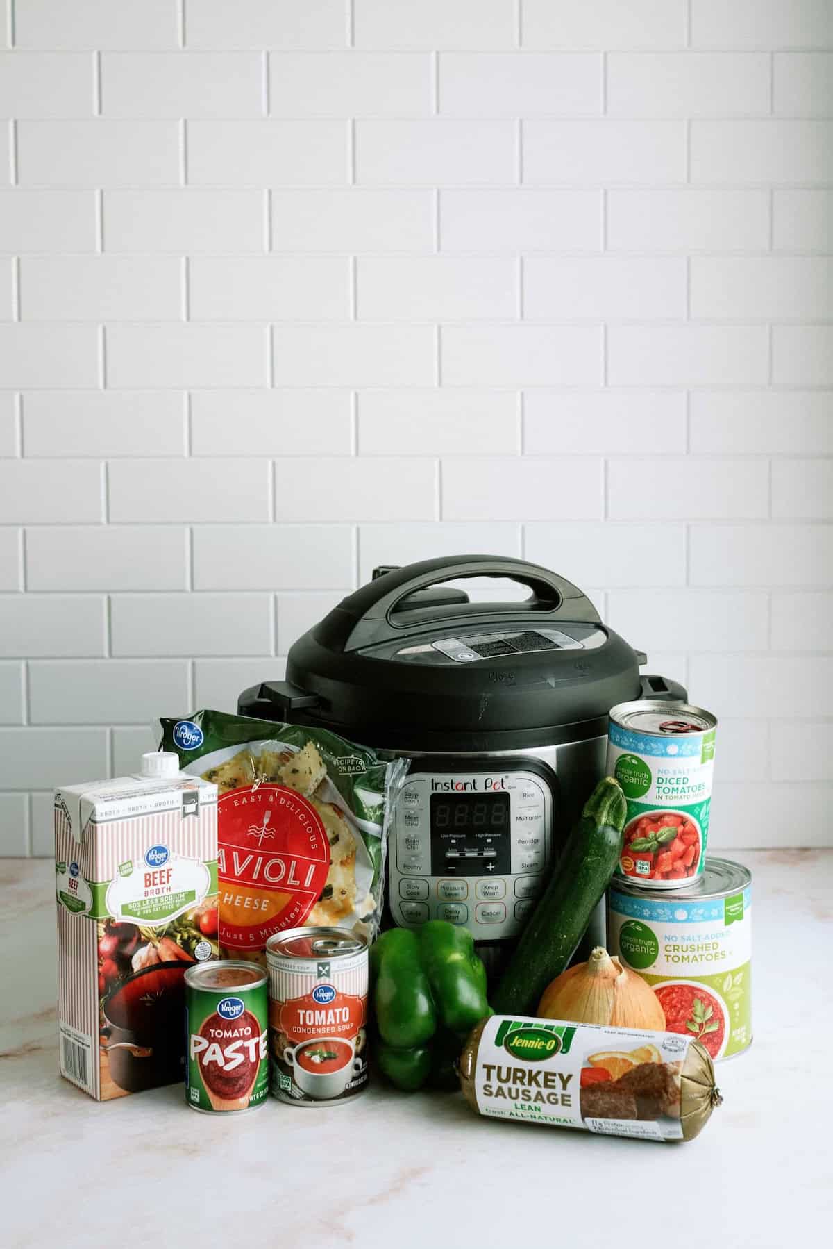 An Instant Pot is surrounded by ingredients including beef broth, ravioli, tomato paste, canned tomatoes, zucchini, green bell pepper, onion, and turkey sausage.