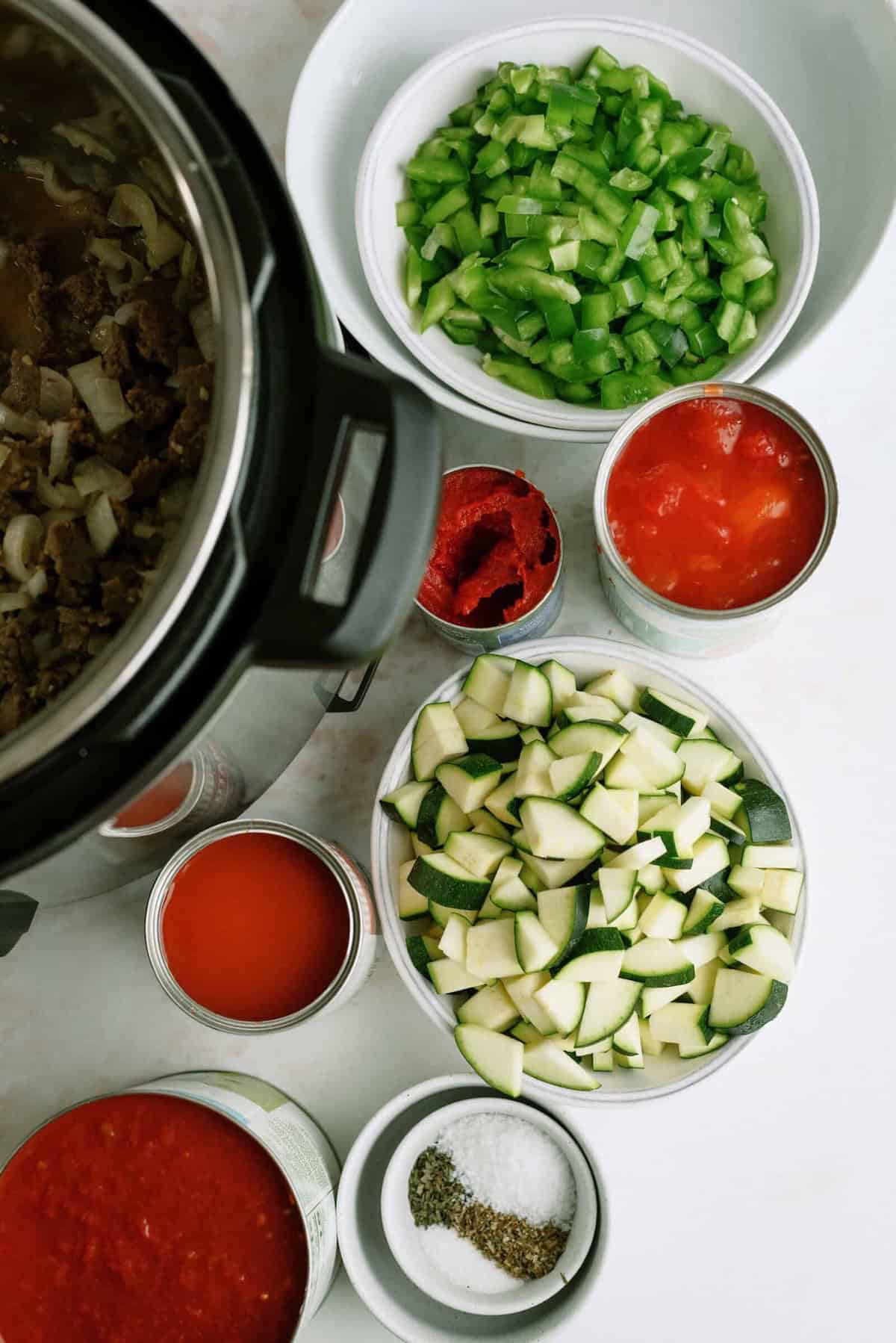 Top-down view of an Instant Pot with bowls of chopped green bell pepper, zucchini, tomato paste, canned tomatoes, tomato sauce, and small bowls of salt and pepper on a white surface.