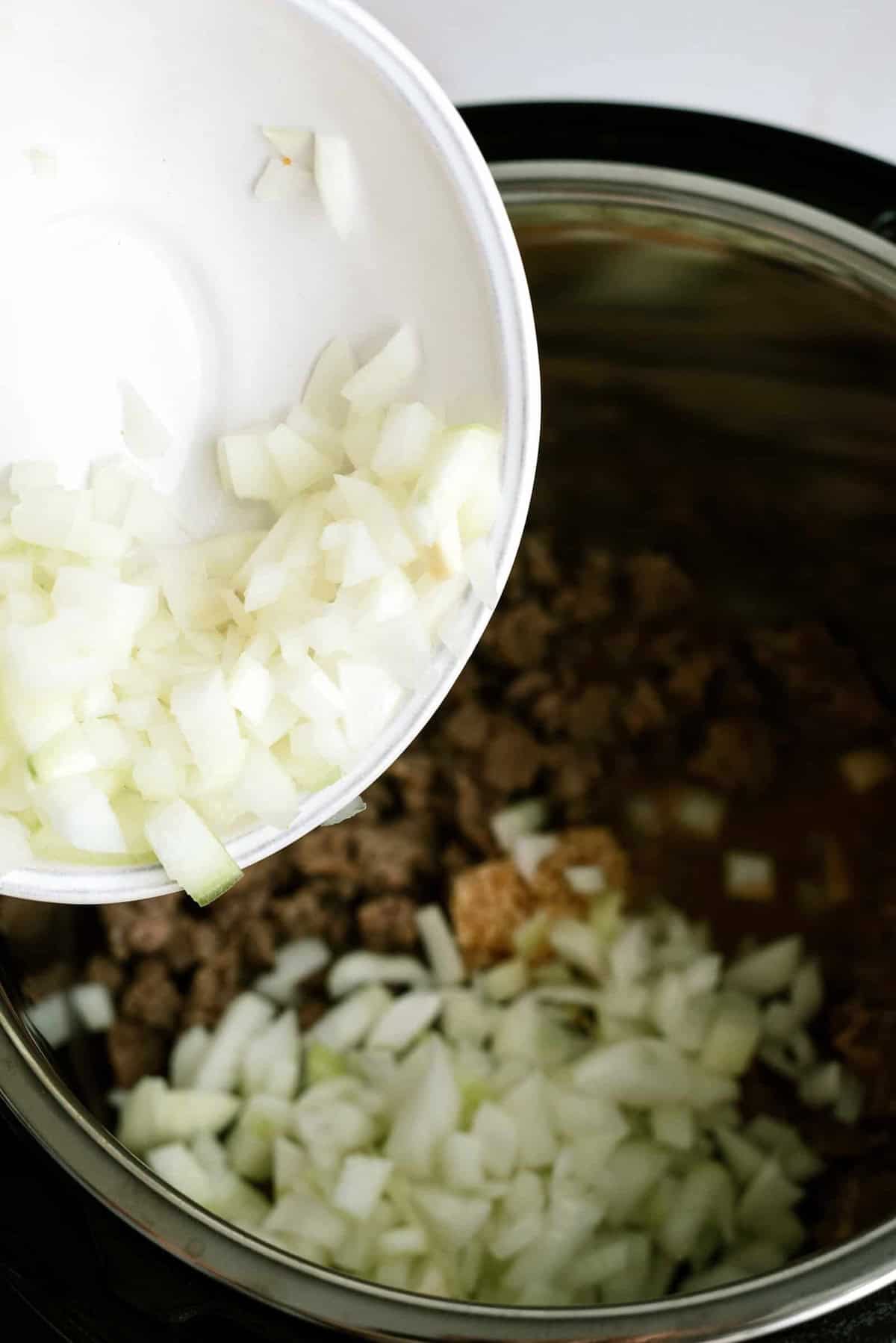 Chopped onions are being poured from a white bowl into a pot with browned ground meat.