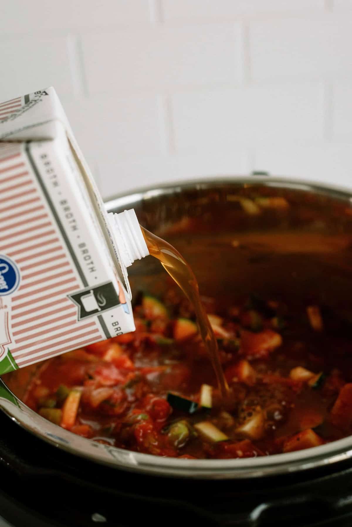 A carton of broth is being poured into a pot containing a tomato-based soup with chopped vegetables.