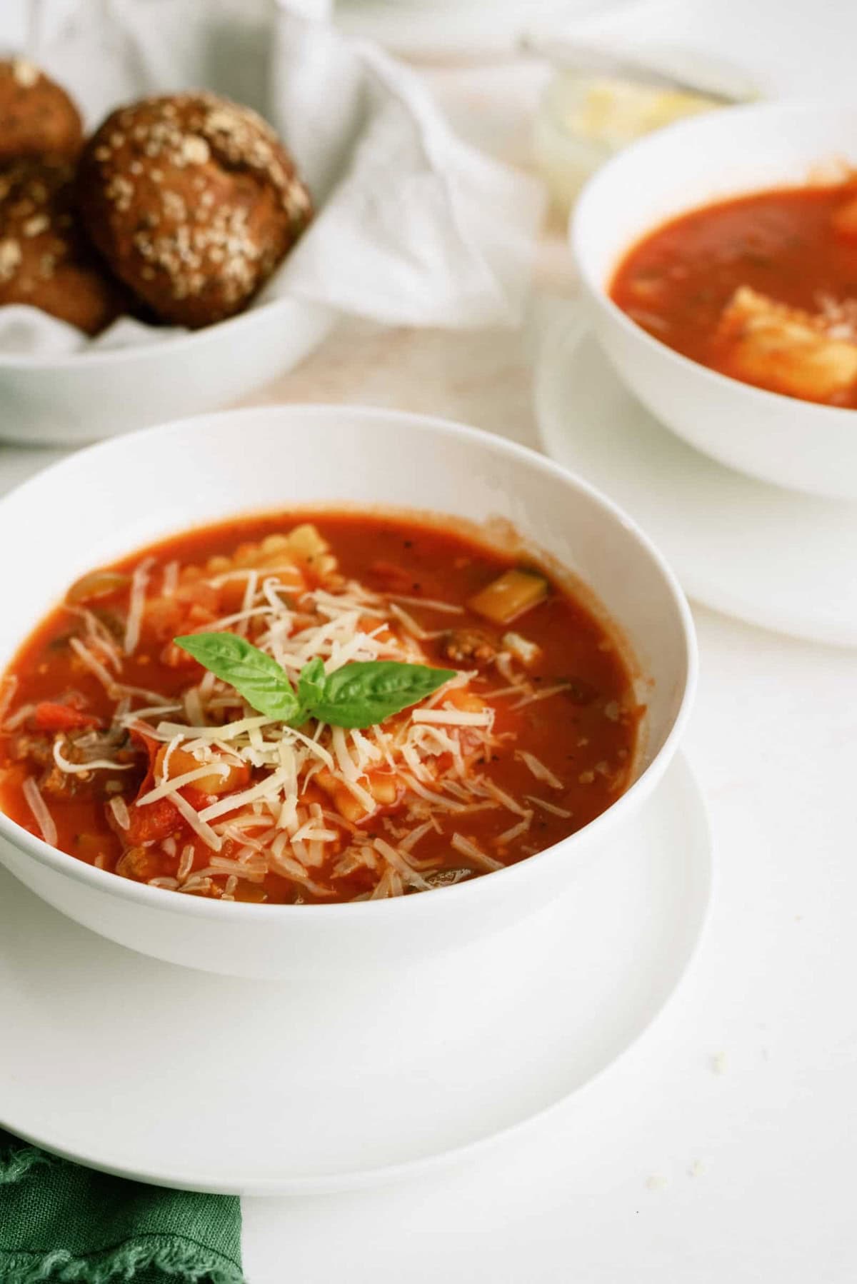 A bowl of vegetable soup topped with grated cheese and a basil leaf, with bread rolls and another bowl of soup in the background.