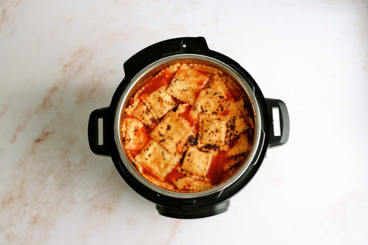 Overhead view of ravioli in tomato sauce with herbs inside an electric pressure cooker on a light marble surface.