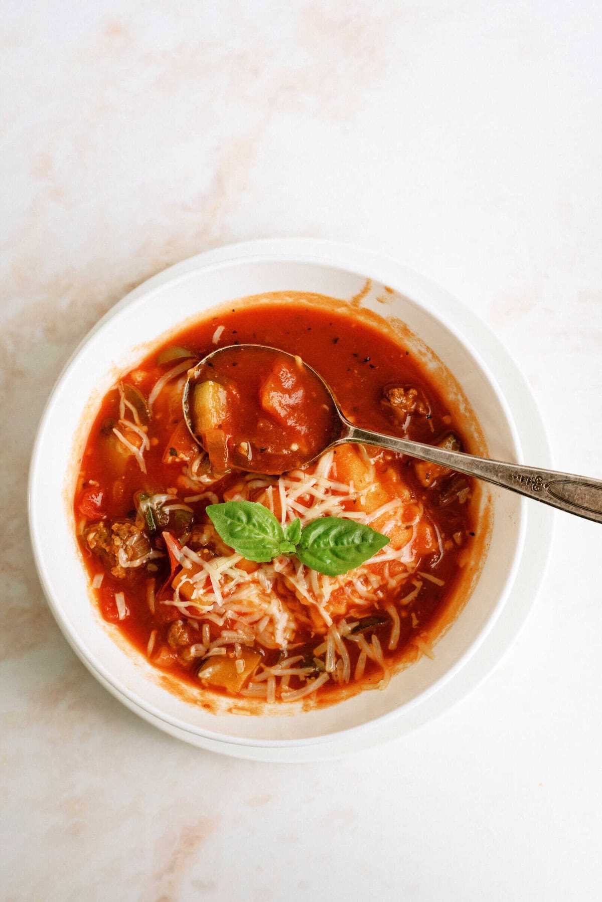 A white bowl of tomato-based soup with pasta, ground meat, grated cheese, and a fresh basil leaf, with a spoon resting inside.