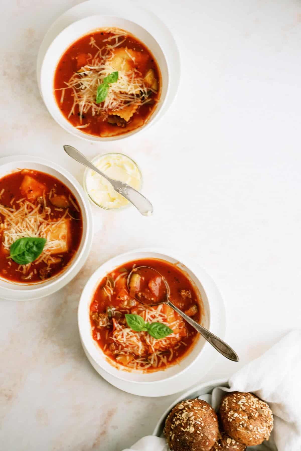 Three bowls of tomato-based soup with grated cheese and basil, a small dish of butter with a knife, and a basket of seeded bread rolls on a white surface.