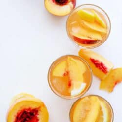 Overhead view of glasses filled with peach slices and liquid, surrounded by fresh sliced peaches on a white surface.