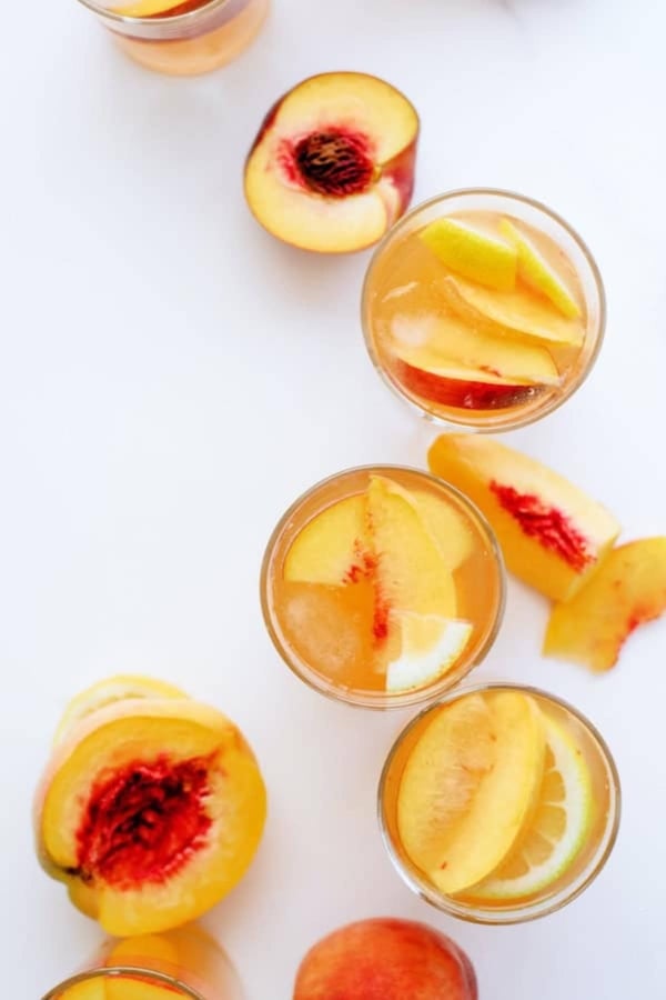 Overhead view of glasses filled with peach slices and liquid, surrounded by fresh sliced peaches on a white surface.