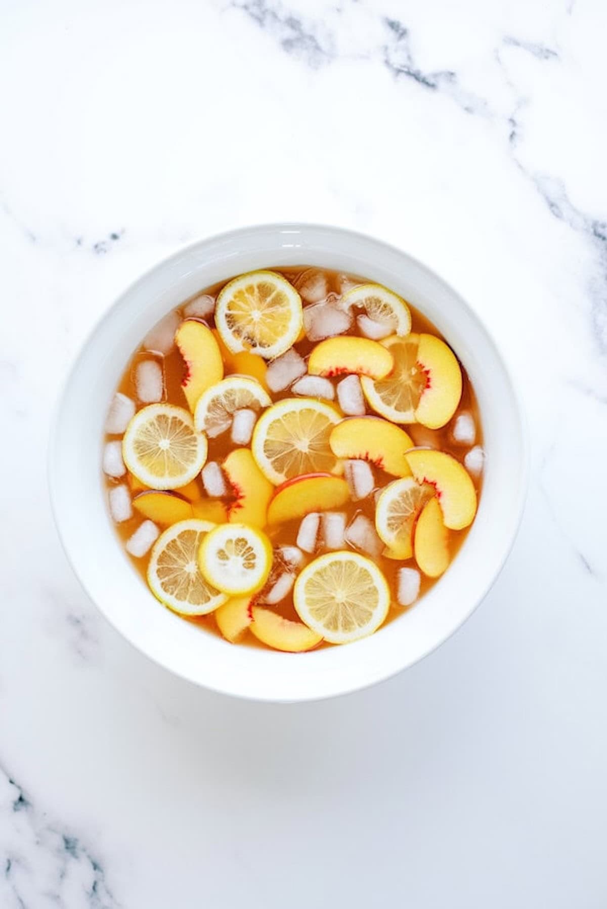 A white bowl filled with iced tea, lemon slices, peach slices, and ice cubes sits on a white marble surface.