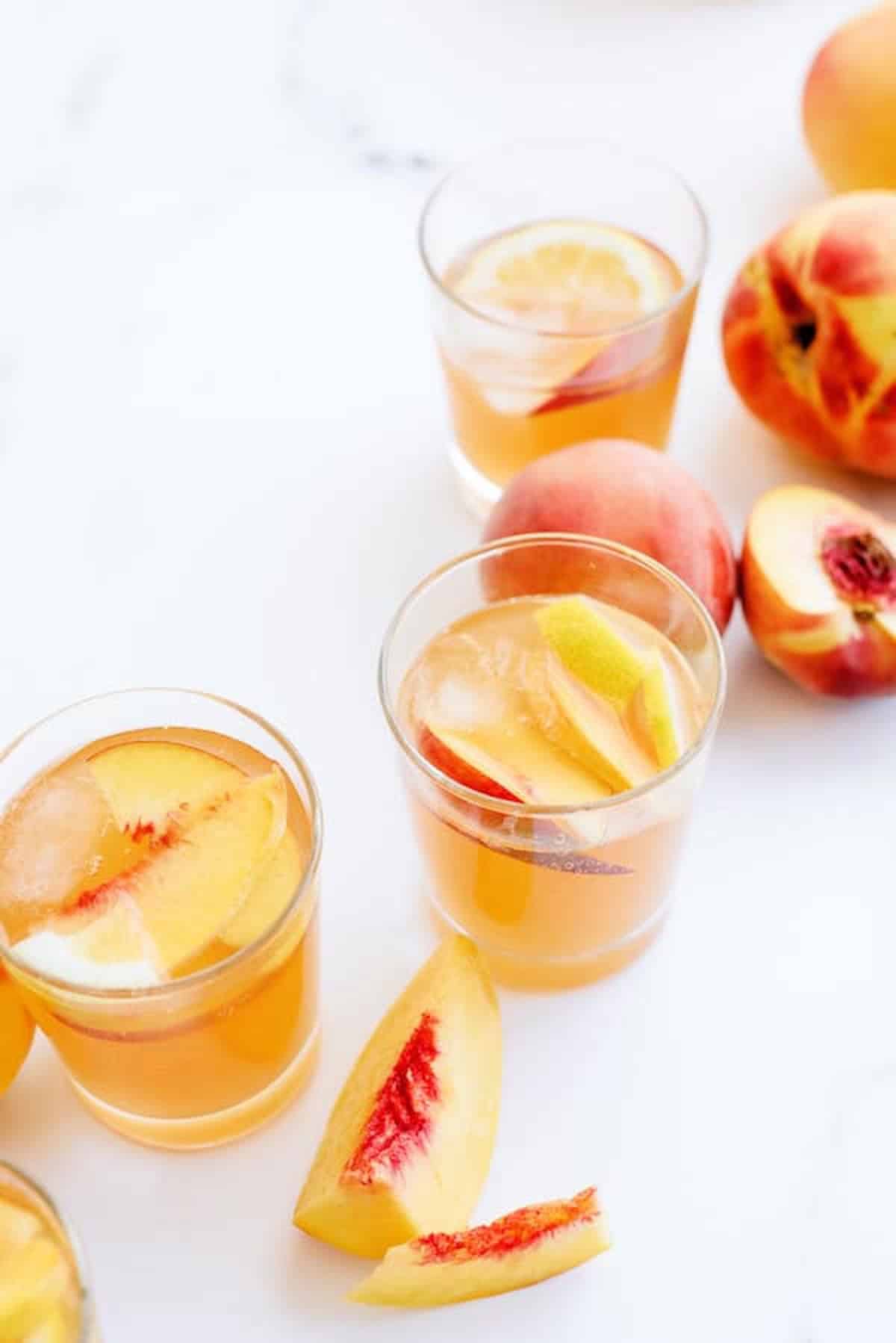 Three glasses filled with iced peach tea are placed on a white surface, surrounded by fresh peaches and peach slices.