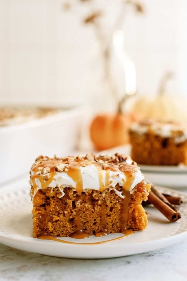 A slice of pumpkin cake topped with frosting and a drizzle of caramel sauce on a white plate, with more cake and a pumpkin in the blurred background.