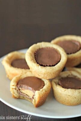 Plate of five peanut butter cup cookies, one with a bite taken out. Cookies have soft, golden-brown edges with whole peanut butter cups in the center.