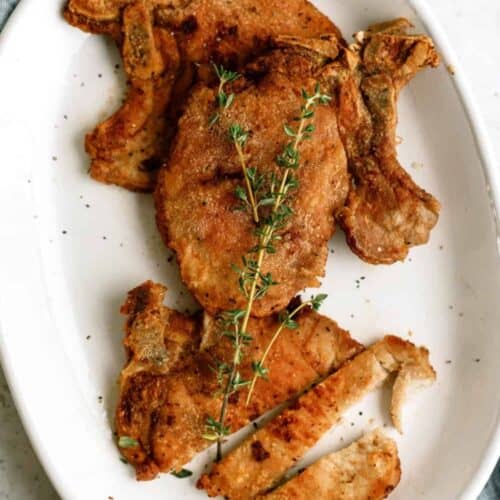 A white oval plate with fried pork chops, one sliced, garnished with a sprig of fresh thyme.