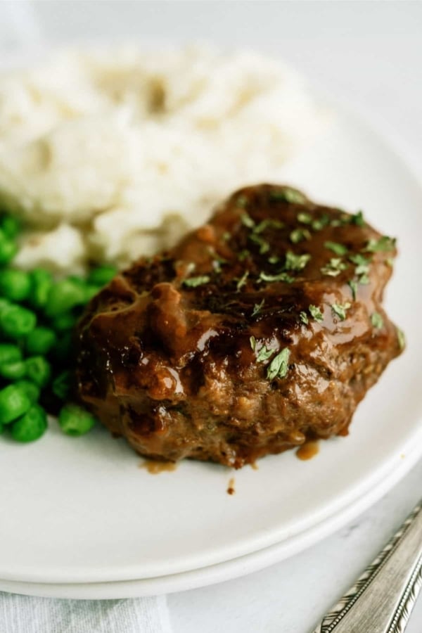 A plate with a cooked hamburger steak topped with gravy, accompanied by a serving of peas and mashed potatoes.