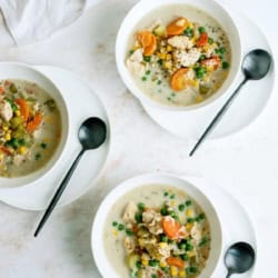 Three white bowls of creamy vegetable and chicken soup with peas, carrots, and corn are on a light surface, each with a black spoon. A plate of bread rolls is in the upper left corner.