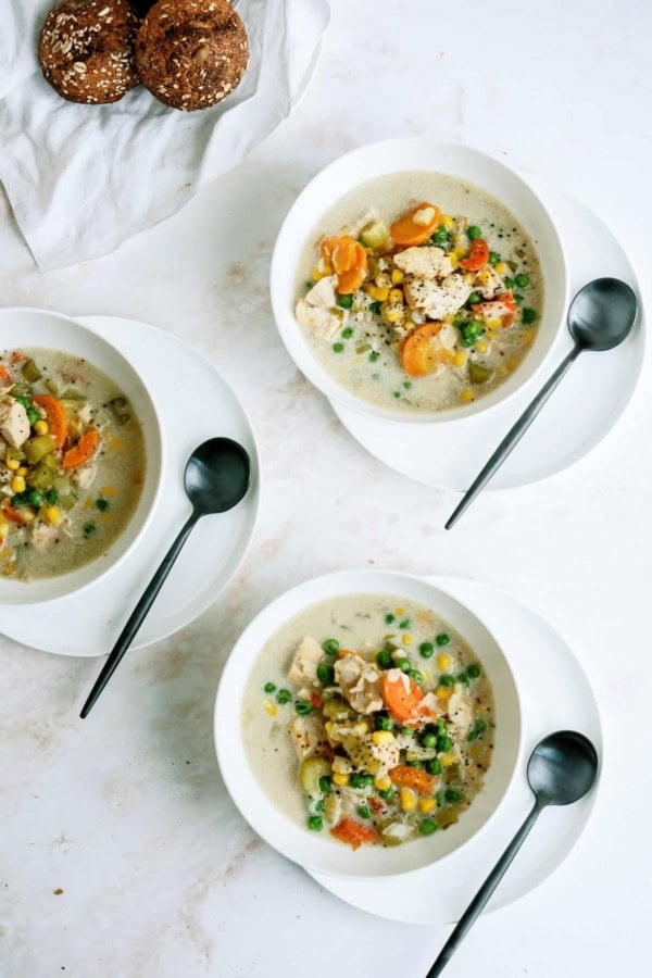 Three white bowls of creamy vegetable and chicken soup with peas, carrots, and corn are on a light surface, each with a black spoon. A plate of bread rolls is in the upper left corner.