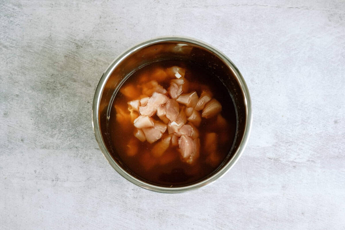 A stainless steel bowl contains raw, diced chicken pieces marinating in a dark brown liquid on a light textured surface.