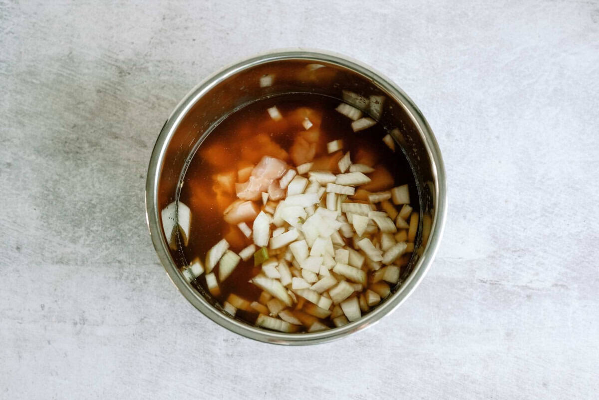 Chopped onions and raw cubed chicken in brown broth inside a stainless steel pot, viewed from above on a light-colored surface.