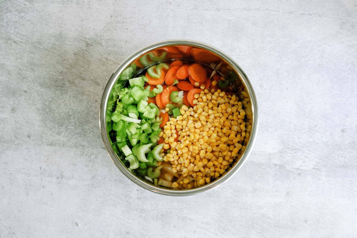A metal bowl filled with chopped celery, sliced carrots, and yellow lentils, placed on a light gray surface.