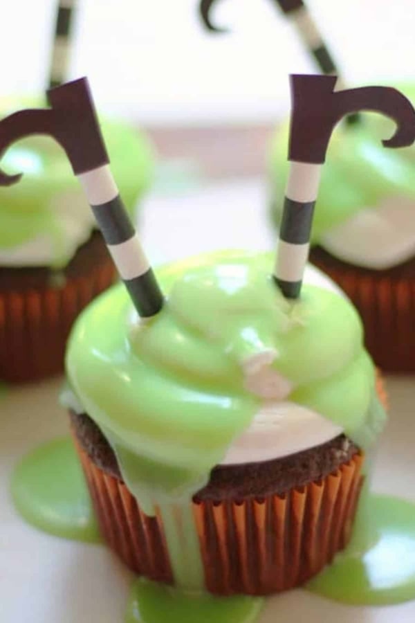 Cupcake with green frosting and two striped paper straws topped with black curled paper cutouts, resembling witches legs.