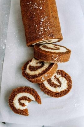 A rolled cake with a brown exterior and white creamy filling, sliced into several pieces, placed on parchment paper with a dusting of powdered sugar.