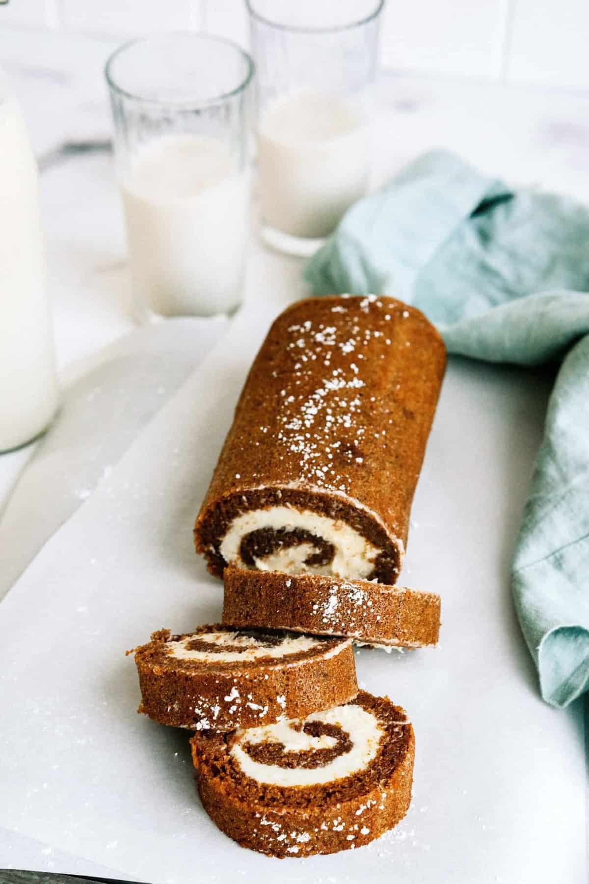 A sliced roll cake with cream filling is placed on a sheet of parchment paper. Two glasses of milk and a light blue cloth are in the background.