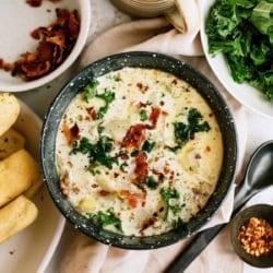 A bowl of creamy soup with chopped greens and bacon bits, next to a plate of breadsticks, a mug, dishes with bacon bits, and a bowl of fresh greens. Spoon and pepper flakes nearby.
