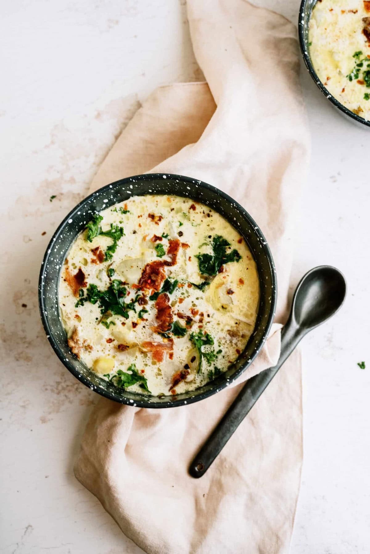 A bowl of creamy soup with visible pieces of bacon, leafy greens, and chopped vegetables, placed on a beige napkin with a spoon beside it.