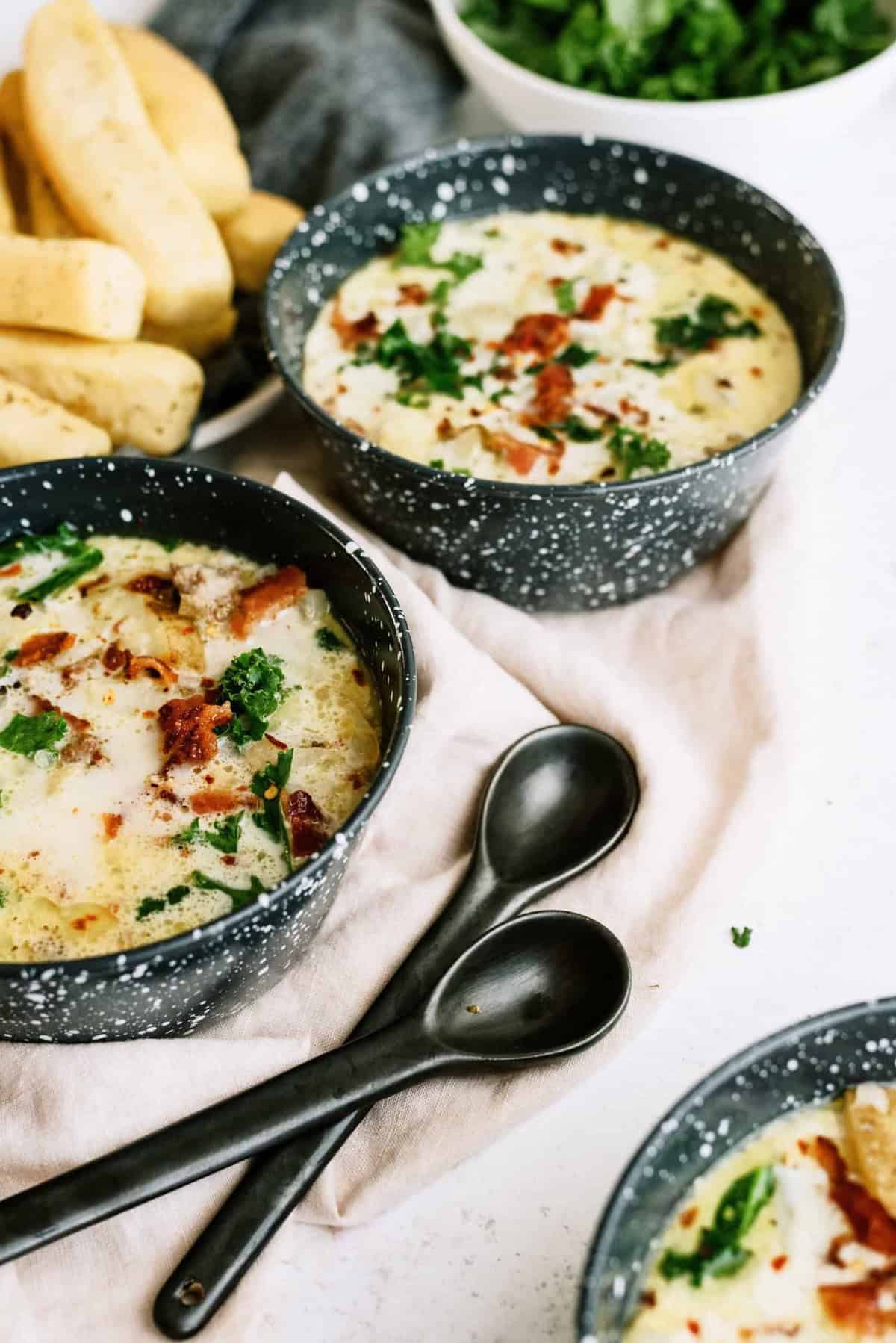 Three bowls of soup with herbs and bacon bits are placed on a light cloth, with two black spoons beside them and breadsticks and salad visible in the background.