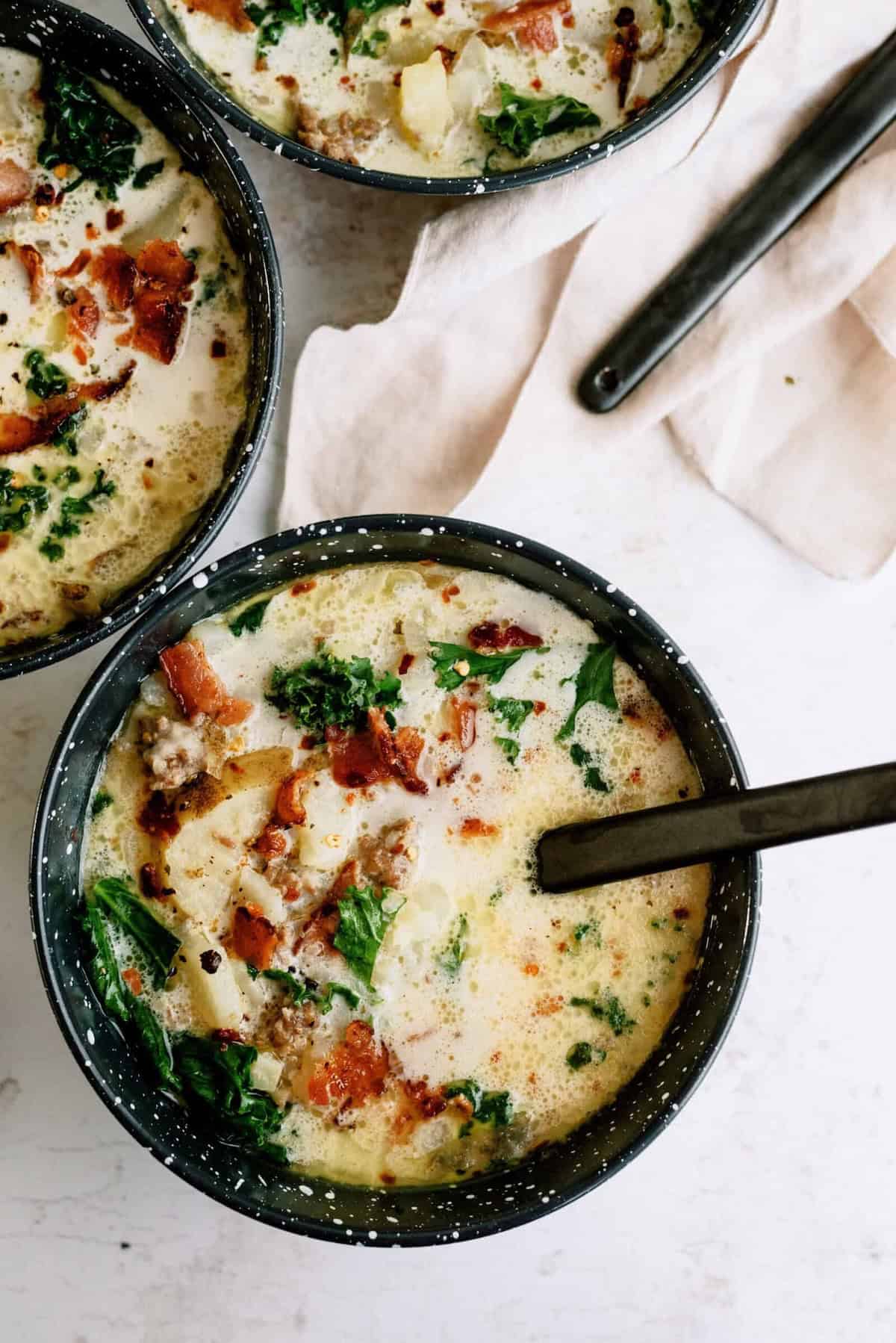 Three bowls of creamy soup with visible chopped bacon, potatoes, greens, and herbs, placed on a light surface with spoons and a beige napkin nearby.