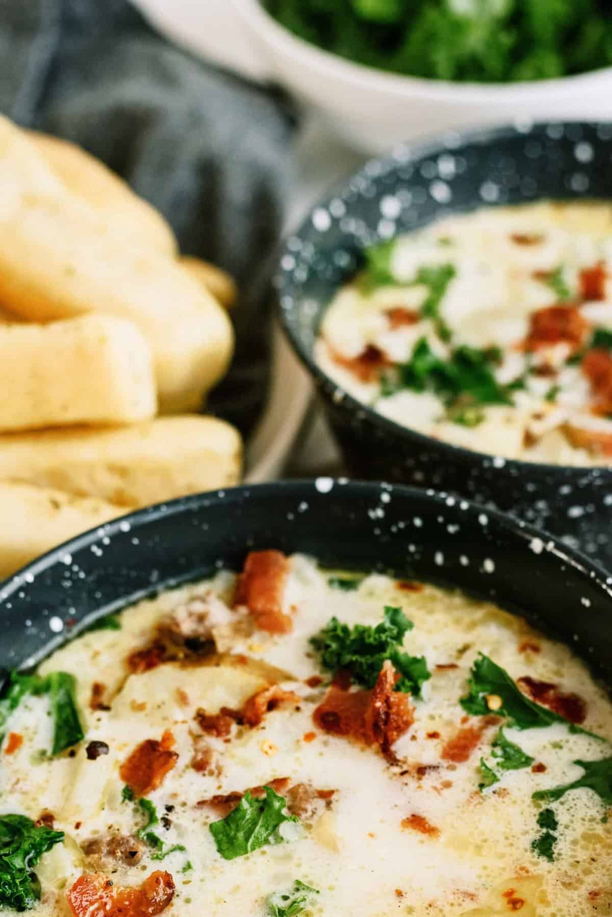Two bowls of creamy soup garnished with bacon and kale, next to a plate of breadsticks.