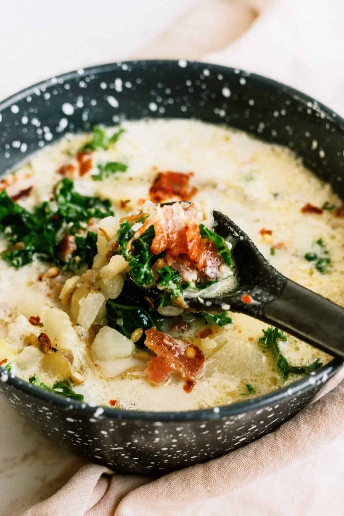 A creamy soup with chunks of potato, kale, and crispy bacon pieces in a black bowl, with a spoon lifting a serving.