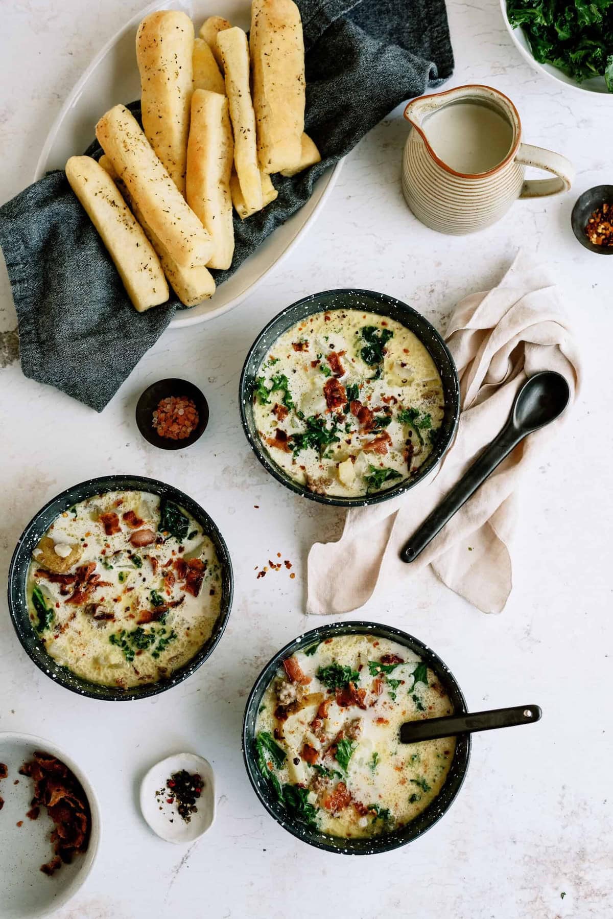 Three bowls of creamy soup with greens and bacon are on a table, accompanied by breadsticks, a small jug, and small dishes of seasonings.