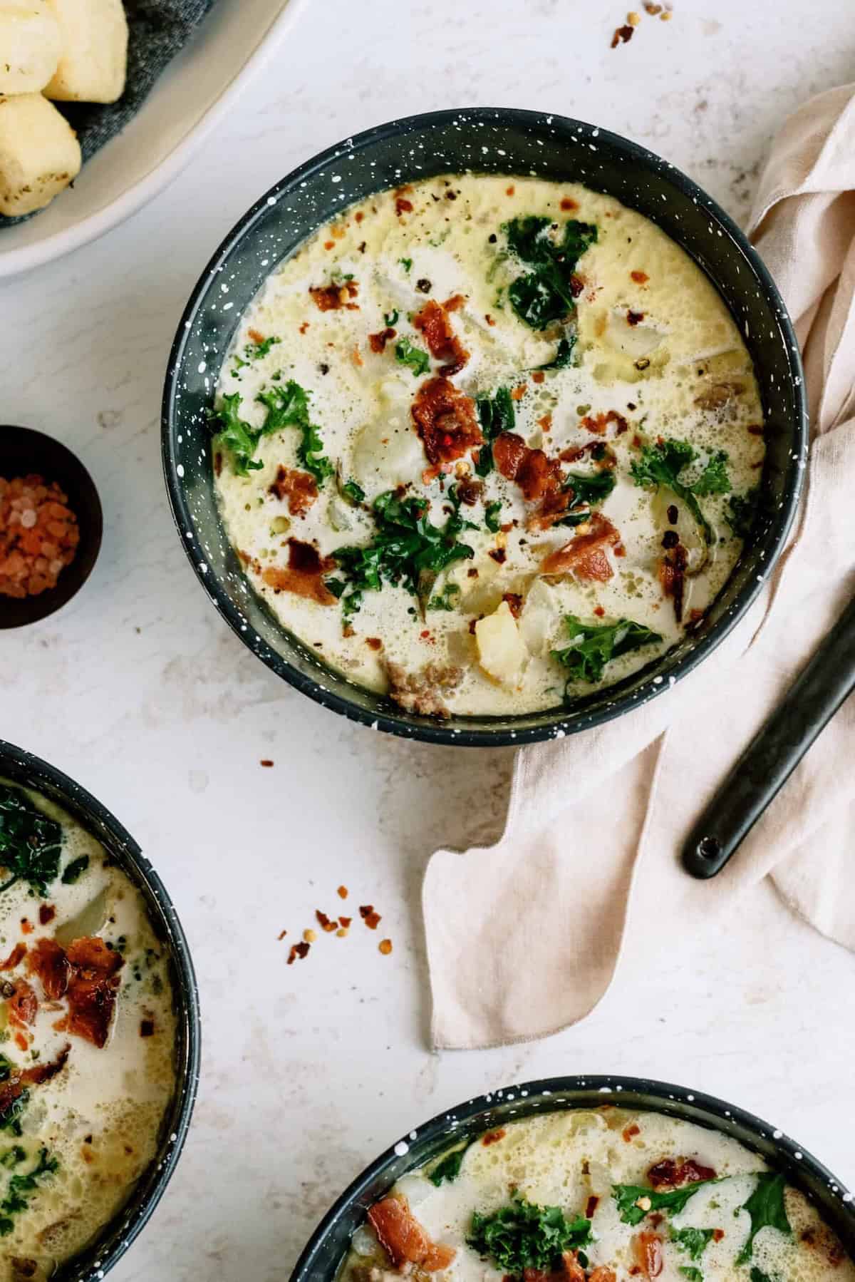 A bowl of creamy soup with potatoes, kale, and bacon pieces, garnished with red pepper flakes, sits on a light surface next to a napkin and spoon.