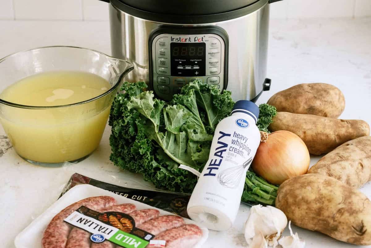 Ingredients for a soup are arranged in front of an Instant Pot, including broth, kale, potatoes, sausage, onion, garlic, and heavy cream.