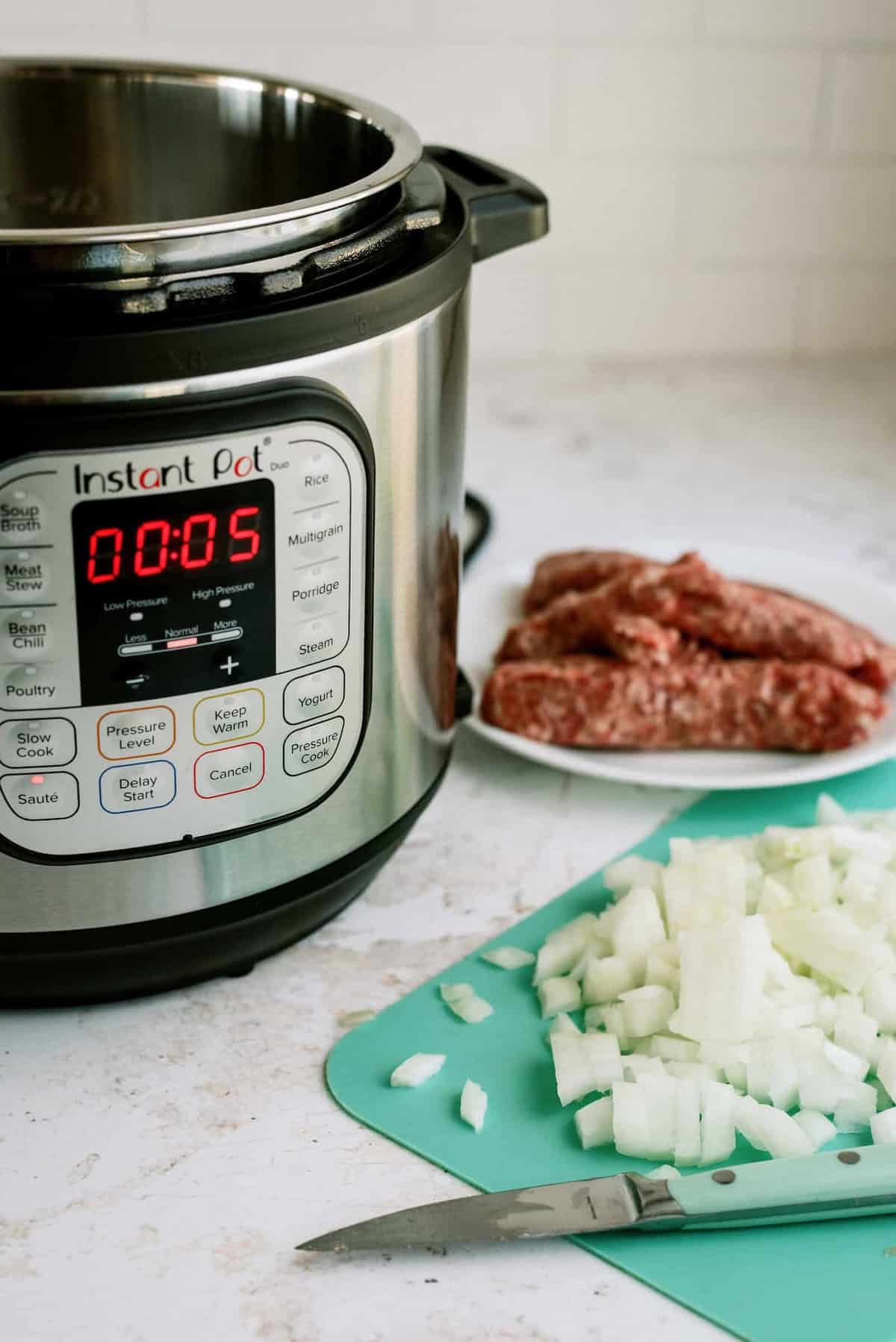 An Instant Pot with a timer set to 5 minutes sits on a counter next to a plate of raw meat and a cutting board with chopped onions and a knife.