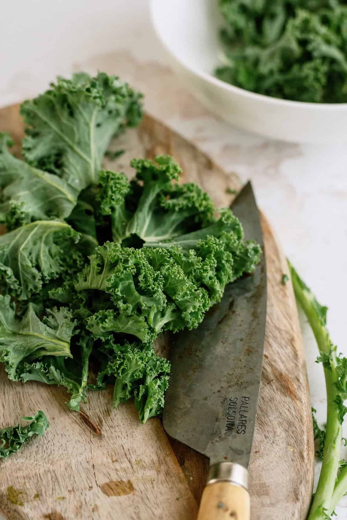 Curly kale leaves on a wooden cutting board with a chef’s knife; a white bowl with more kale is in the background.