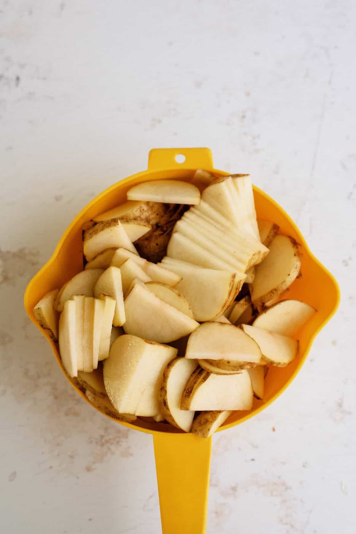 Sliced potatoes with skin in a yellow colander on a light-colored surface.