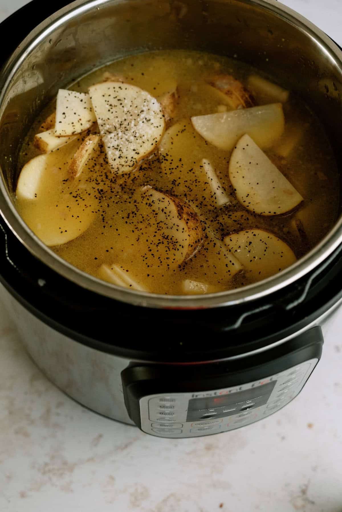 A pressure cooker filled with broth, potato slices, and ground black pepper, sits on a light countertop.