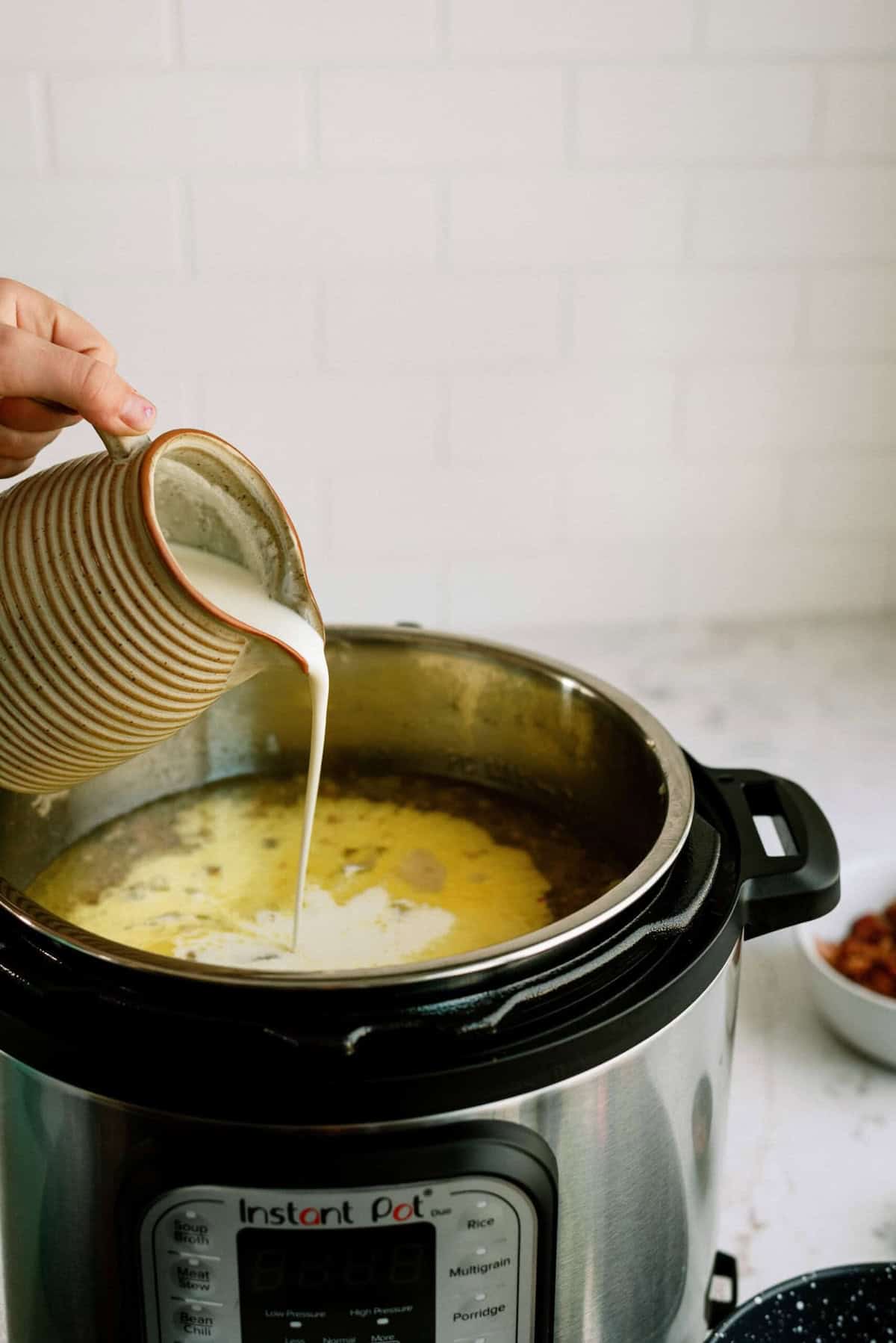 A hand pours cream from a striped ceramic cup into a soup cooking in an open Instant Pot on a kitchen counter.