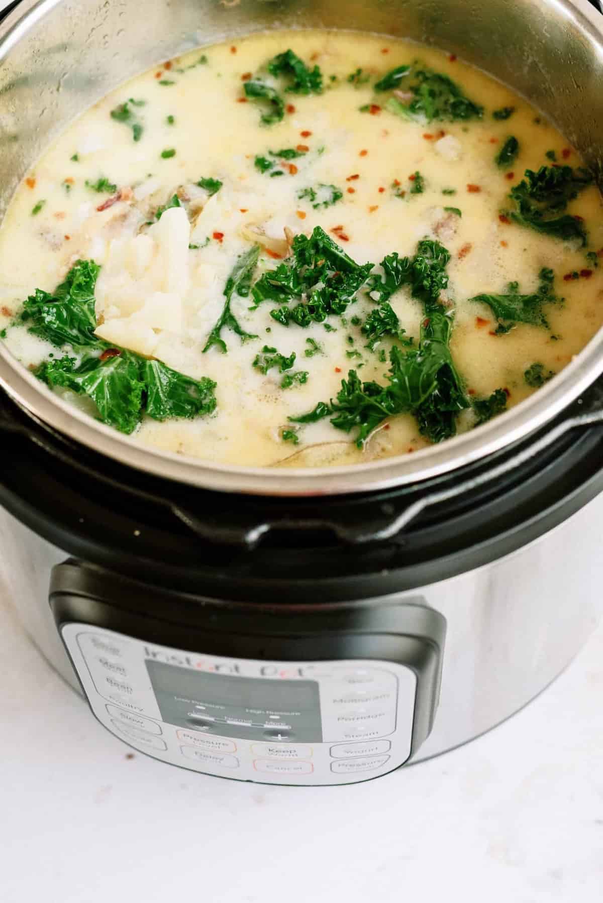 An Instant Pot filled with creamy soup containing pieces of kale and what appears to be chicken or fish, with red pepper flakes sprinkled on top.