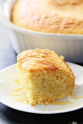 A slice of cornbread on a white plate with the remaining cornbread in a baking dish in the background.