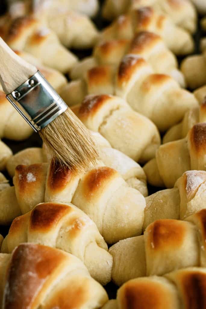A pastry brush, brushing honey butter on baked Lion House Rolls.
