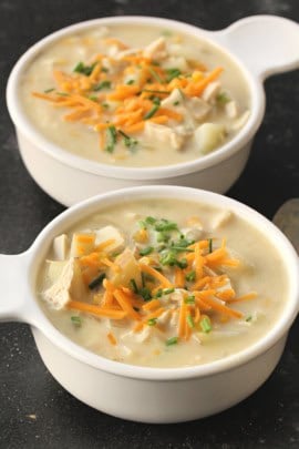 Two white bowls of creamy turkey soup topped with shredded cheese and chopped chives on a dark surface.